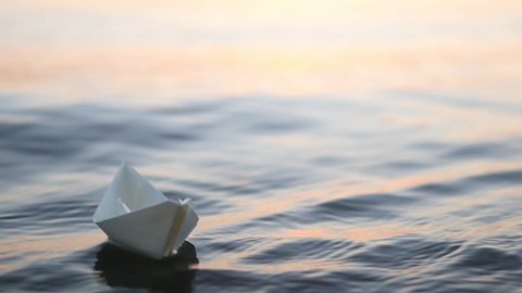 A paper boat is floating on the waves in the water at a beautiful sunset. Origami ship Sailing. The concept of a dream, future, childhood, freedom or hope. Slow motion