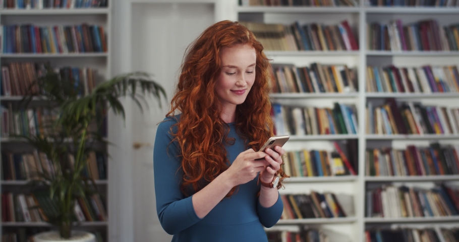 Red-haired young attractive Caucasian woman tapping and typing on the smartphone and then making surprised face and saying wow to the camera while standing in the library.