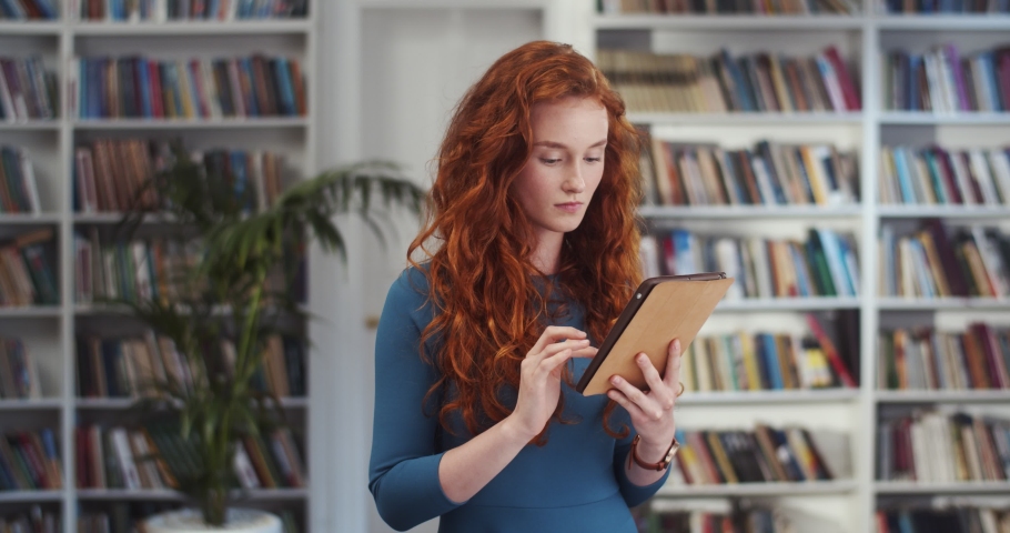 Young Caucasian pretty woman tapping, typing and scrolling on the tablet device in the library and then smiling happily to the camera. Portrait shot.