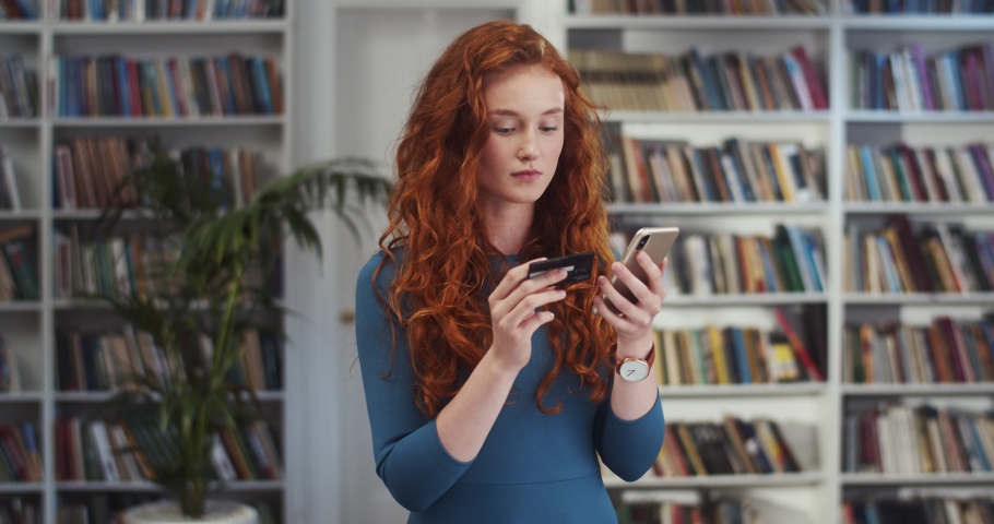 Caucasian young pretty red-haired woman tapping while shopping online with a credit card and smiling in the library.