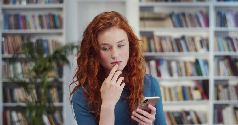 Young Caucasian beautiful woman with red hair standing in the library, thinking and tapping or texting on the smartphone.