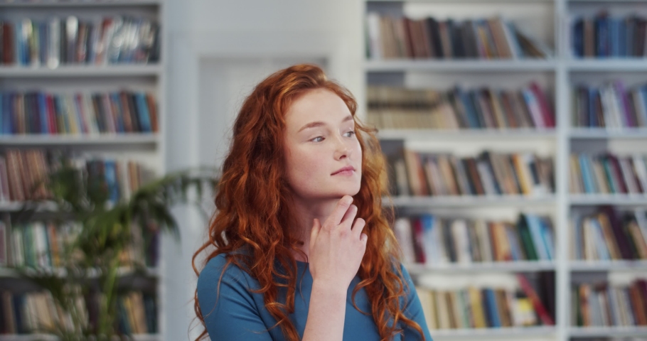 Young Caucasian pretty woman with red hair standing in the library and dreaming while thinking deeply in the library. Portrait.