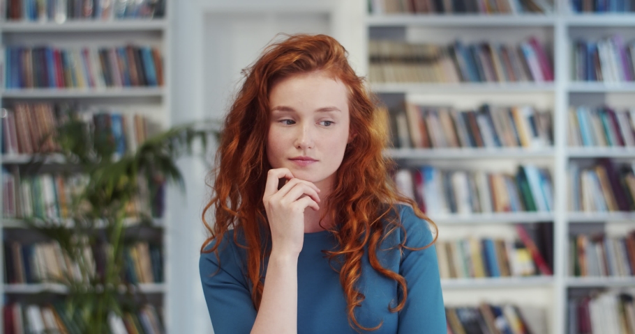 Portrait shot of the red-haired Caucasian young and pretty girl thinking like choosing something in the library.