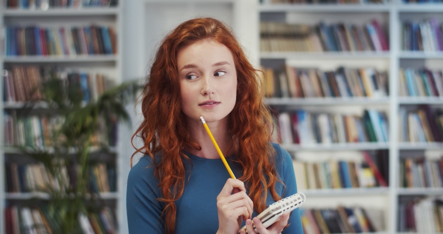 Caucasian attractive young girl with long red hair standing in the library with pencil and notebook while thinking, smiling while noting something in the library. Portrait shot.
