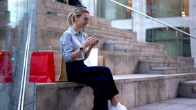 Carefree woman in spectacles sitting outdoors and checking purchase sale from shopping bag for typing sms message to friend and sharing about Black Friday benefits, millennial girl chatting via mobile