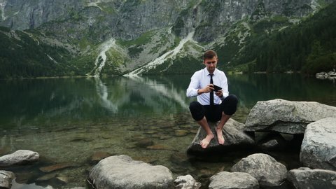 Gambling businessman playing a game on mobile phone, sitting on a stone near a lake in the rocky mountains, win the game, rejoices in victory
