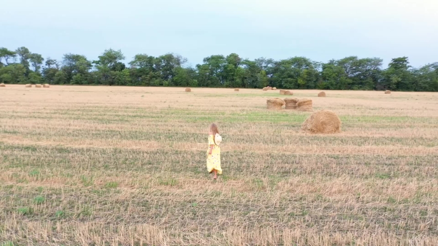Girl with long hair in dress is  running across an agricultural field with haystack