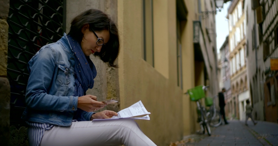 woman is examining papers sitting in small narrow lane, holding smartphone in hands