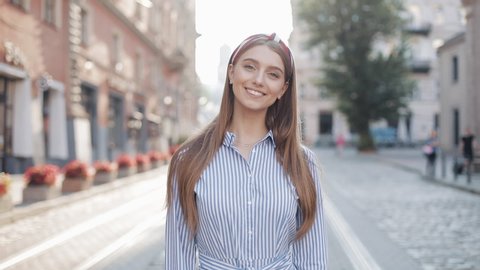 Portrait of Young Happy Charming Girl with Straight Brown Hair and Blue Eyes Wearing in Striped Dress and Fashionable Headband Smiling Looking to Camera Standing at the City Street Zoom