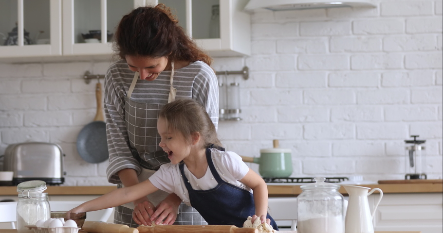 Loving happy mom teaching cute kid daughter learning kneading dough with rolling pin, funny child girl helping mother preparing cookies cooking baking pastry playing in modern kitchen at home Royalty-Free Stock Footage #1037333450
