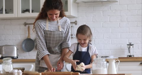 Loving happy mom teaching cute kid daughter learning kneading dough with rolling pin, funny child girl helping mother preparing cookies cooking baking pastry playing in modern kitchen at home