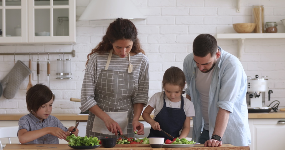Happy family mom dad and kids siblings cooking together, young parents teaching children son daughter learning cutting fresh vegetable salad prepare healthy food in modern kitchen interior together | Shutterstock HD Video #1037333453