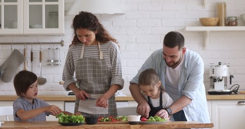 Happy family mom dad and kids siblings cooking together, young parents teaching children son daughter learning cutting fresh vegetable salad prepare healthy food in modern kitchen interior together