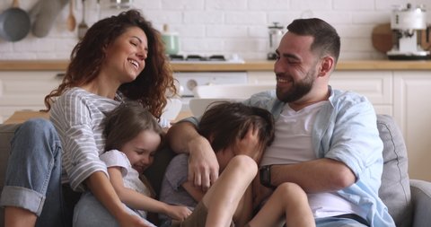 Happy family or four mum dad with cute little kids son daughter tickling having fun relax on sofa together, young carefree parents and children laugh play cuddling at home enjoy funny game on couch