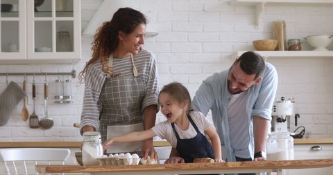 Happy family mom dad and kid daughter kneading dough baking pastry play with flour cooking together, young parents teaching child girl learning prepare cookies laughing having fun in modern kitchen