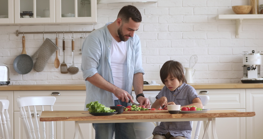 School boy son helping young happy father cutting fresh vegetables for healthy salad in modern kitchen interior, smiling dad teaching child learning cooking preparing dinner meal together at home Royalty-Free Stock Footage #1037333468
