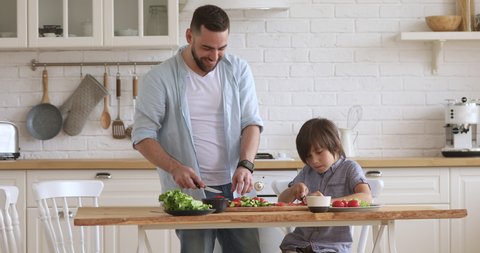School boy son helping young happy father cutting fresh vegetables for healthy salad in modern kitchen interior, smiling dad teaching child learning cooking preparing dinner meal together at home