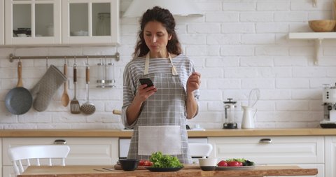 Happy young woman preparing vegetable meal searching checking online vegan recipe in smartphone, smiling vegetarian girl cooking healthy food salad using mobile cookbook menu apps in modern kitchen