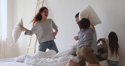 Happy playful family of four parents and little cute kids having fun pillow fight on bed, carefree couple with small funny small children laughing playing leisure game in bedroom on weekend together