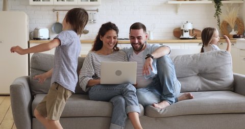 Happy family or four enjoy weekend lifestyle relaxing in modern kitchen interior, young parents couple sit on couch using laptop funny little cute active kids children running playing in modern home