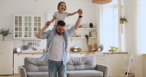 Loving young dad carrying cute little kid daughter giving piggyback ride at home, happy funny small girl sitting on father shoulder playing carrying child bonding having fun laughing in kitchen room