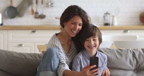 Happy young mother and school son laughing using smartphone funny app take selfie for social media at home, smiling mum having fun with child boy holding cellphone look at screen bonding sit in sofa