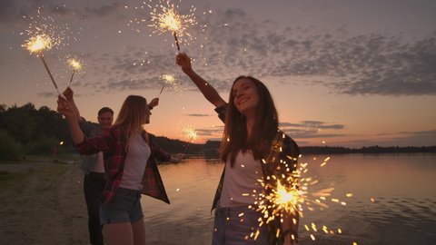 Cheerful male and female friends are running along the beach at sunset, holding sparkling fireworks and runaway lights in slow motion. Dancing and sunset party on the beach.