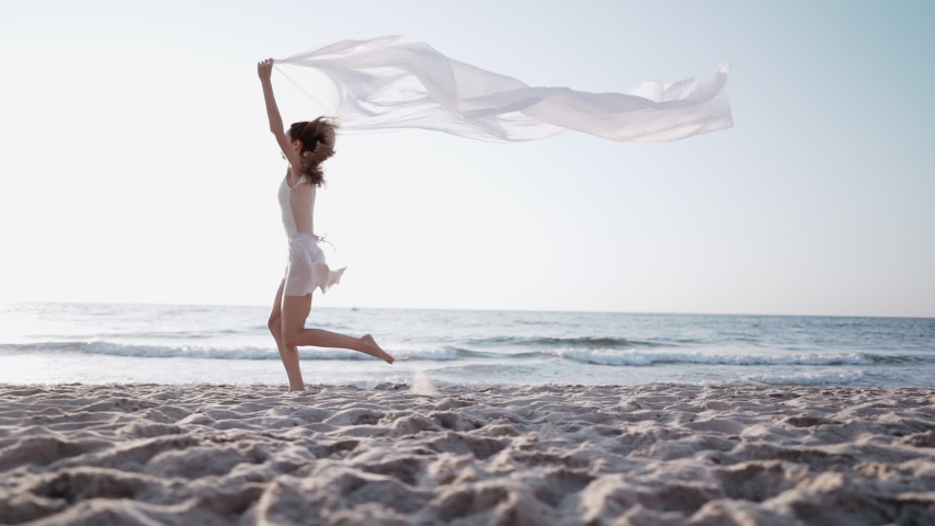 Slim beautiful woman running on sea beach with white silk fabric fluttering in wind.?oncept of tenderness, lightness, art and talent in nature