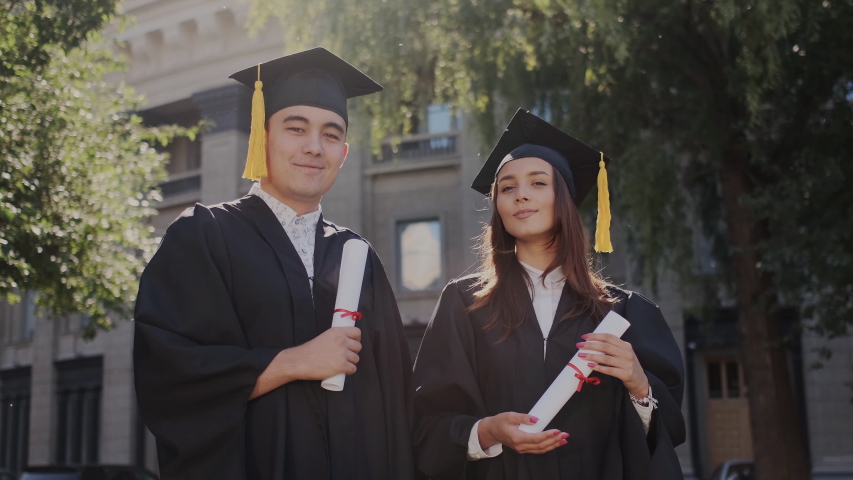 Graduates or students, who successfully finish study. Young multi ethnic guy and girl in academic hat with diploma looking into the camera. Ambitious and proud people in black mantle or gown. Royalty-Free Stock Footage #1037347052