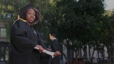 Portrait of the African American happy young graduated woman. She having fun, dancing and making YES gesture. University and Graduates with on the background. Slow motion.