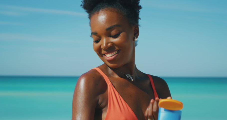 Slow motion of happy young african woman is applying a sunscreen or sun tanning lotion to take care of her skin during a vacation on a beach and smiling in camera. Royalty-Free Stock Footage #1037347928