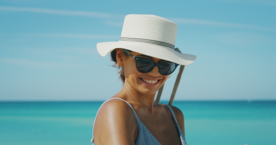Slow motion of happy young brunette woman with hat and sunglasses is applying a sunscreen or sun tanning lotion to take care of her skin during a vacation on a beach and smiling in camera. Royalty-Free Stock Footage #1037348756