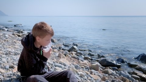 Sick child boy uses nebulizer sits on seaside throw stones. Inhaling inhaler mask. Fibrosis cystic copd and treatment. Asthma pulmonary respiratory breath problem cure. Painkiller sedative gas oxygen.