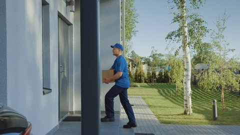Delivery Man Holding Card Board Package Enters Through the Gates and Walks to the House and Knocks. Delivering Postal Parcel. In the Background Beautiful Suburban Neighbourhood. Following Side View