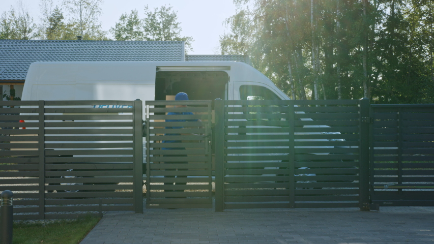 Delivery Man Holding Card Board Package Enters Through the Gates Delivering Postal Parcel. In the Background Delivery Van in the Driveway of Suburban Street