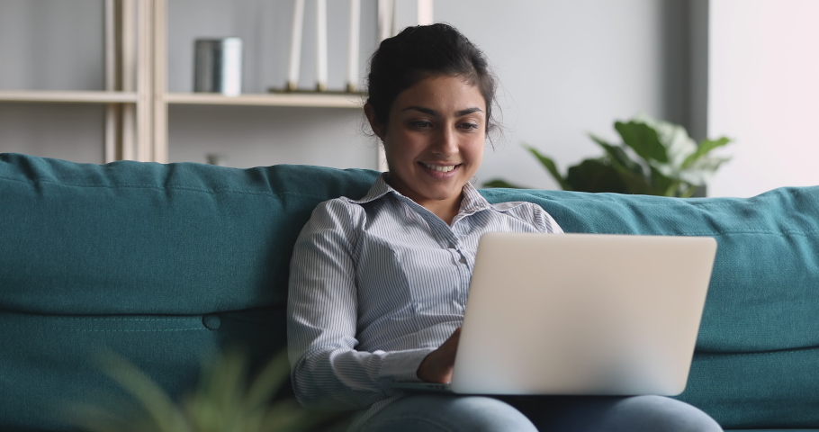 Smiling young indian woman using laptop notebook sitting on couch at home, happy hindu girl typing on computer browsing internet working studying communicating online surfing web looking at screen Royalty-Free Stock Footage #1037352953