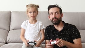 father and young child are playing video game on couch at home, pressing buttons on joystick