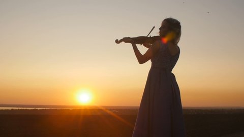 silhouette of a young woman figure in the sunrays playing the violin, girl relaxing and moving bow along the strings of a musical instrument at sunset in a field, concept hobby
