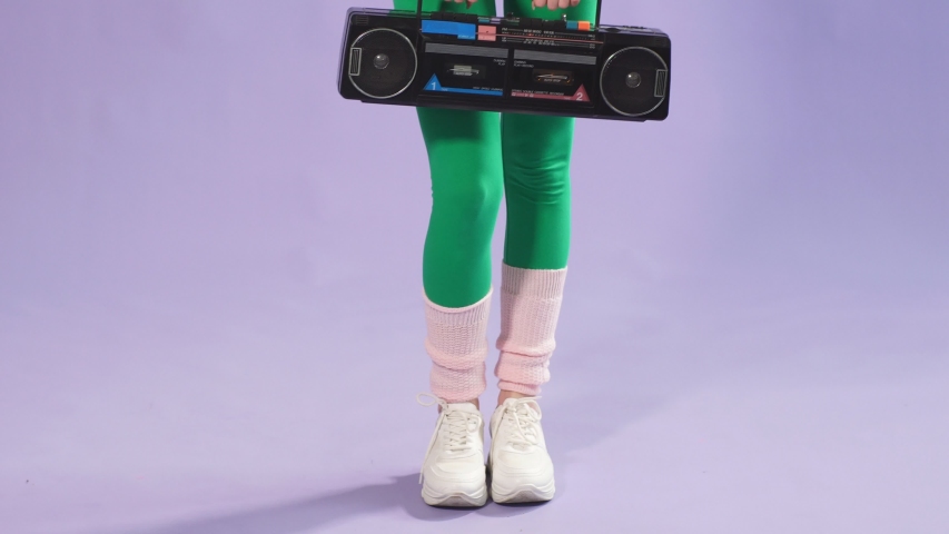 Female aerobics trainer stands with hands forward holding portable stereo cassette player, smiles joyfully in camera. Studio shot over purple background.