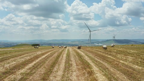 Aerial shot flying over hay field with hay bales below and wind turbines on the background. Landscape of Basilicata in Southern Italy.