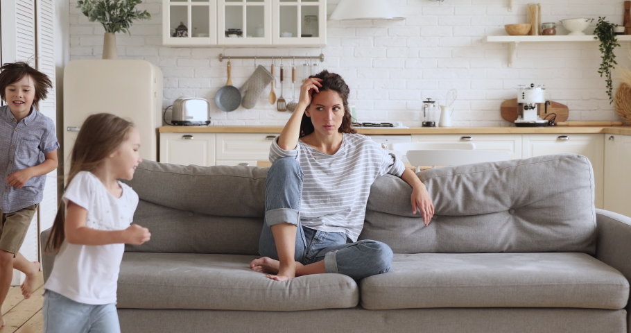 Tired upset young mother sit on sofa feel frustrated about active noisy kids running playing in kitchen, stressed exhausted fatigued single mom annoyed disturbed about disobedient difficult children Royalty-Free Stock Footage #1037398691