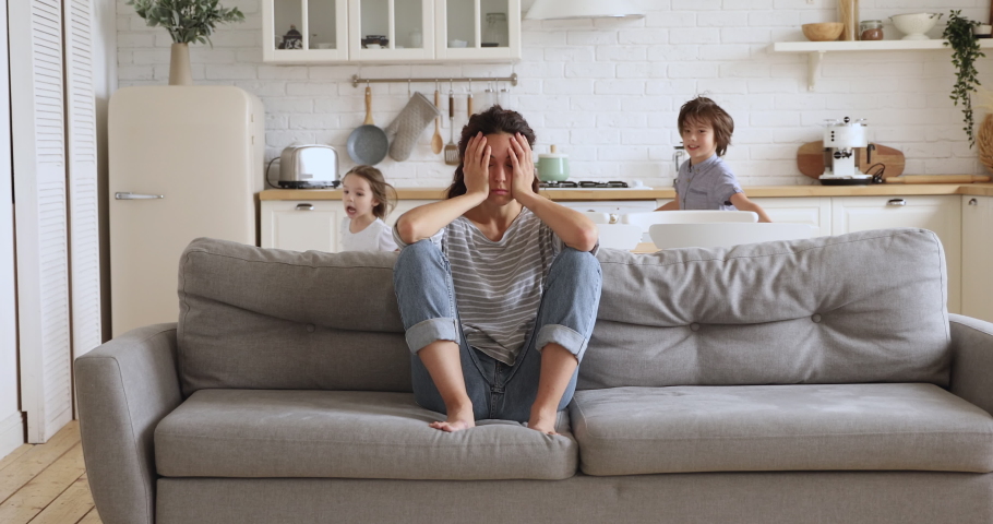 Tired upset young mother sit on sofa feel frustrated about active noisy kids running playing in kitchen, stressed exhausted fatigued single mom annoyed disturbed about disobedient difficult children Royalty-Free Stock Footage #1037398691