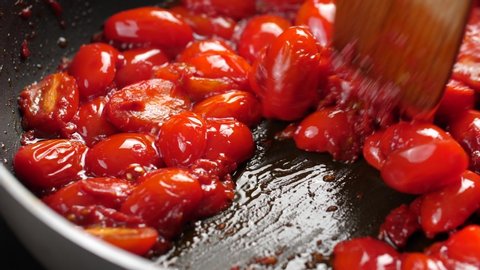 Stirring sauteed halved cherry tomatoes on hot pan using wooden spatula; close up shot in slow motion