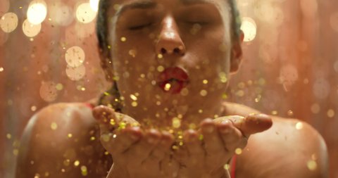 Slow motion front view close up of a young Caucasian woman blowing golden glitter from her hands at the camera, in front of a curtain of shiny silver fringes