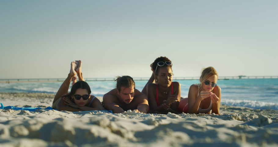 Slow motion of young multi-ethnic friends in swimsuits are lying on a beach enjoying their summer vacation together on a beach with a sea in a sunny day.