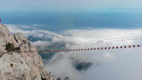 Person walking rope bridge, aerial view. Crossing over a suspension bridge in the mountains, walk above clouds. Extreme sport, risk, overtake, overcome, fear of height. Drone flight