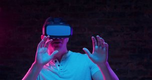 Front view close up of a young Caucasian man wearing a VR headset, smiling and looking around and moving, with both hands raised in front of him as if avoiding things, lit with pink and blue light on