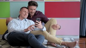 Young gay male couple using phone on arm chair. Video call to friend. Happy gay friendship, relationship, pride concept.
