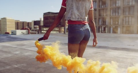Back view close up of a hip young mixed race woman walking and using smoke grenade on an urban rooftop with buildings in the background in slow motion