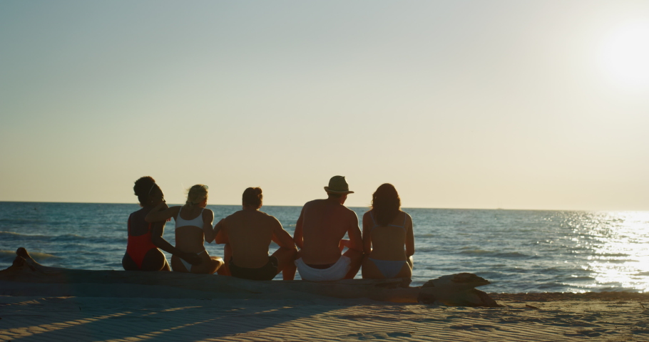 Slow motion of young multi-ethnic friends in swimwear are cheering for a start of their summer vacation together on a beach with a sea on a sunset.